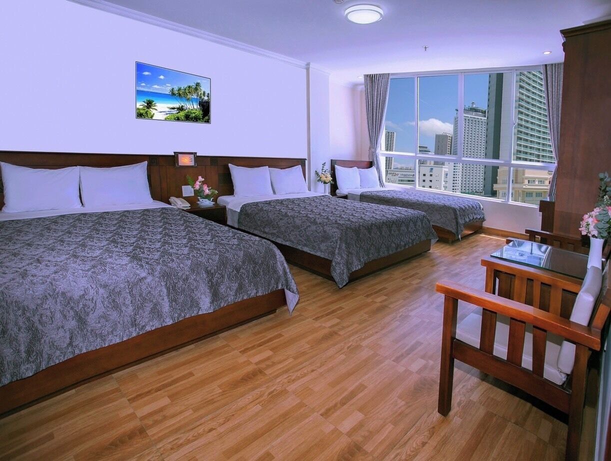 A hotel room with two beds, a wooden floor, a desk with a chair, and a large window with a city view.