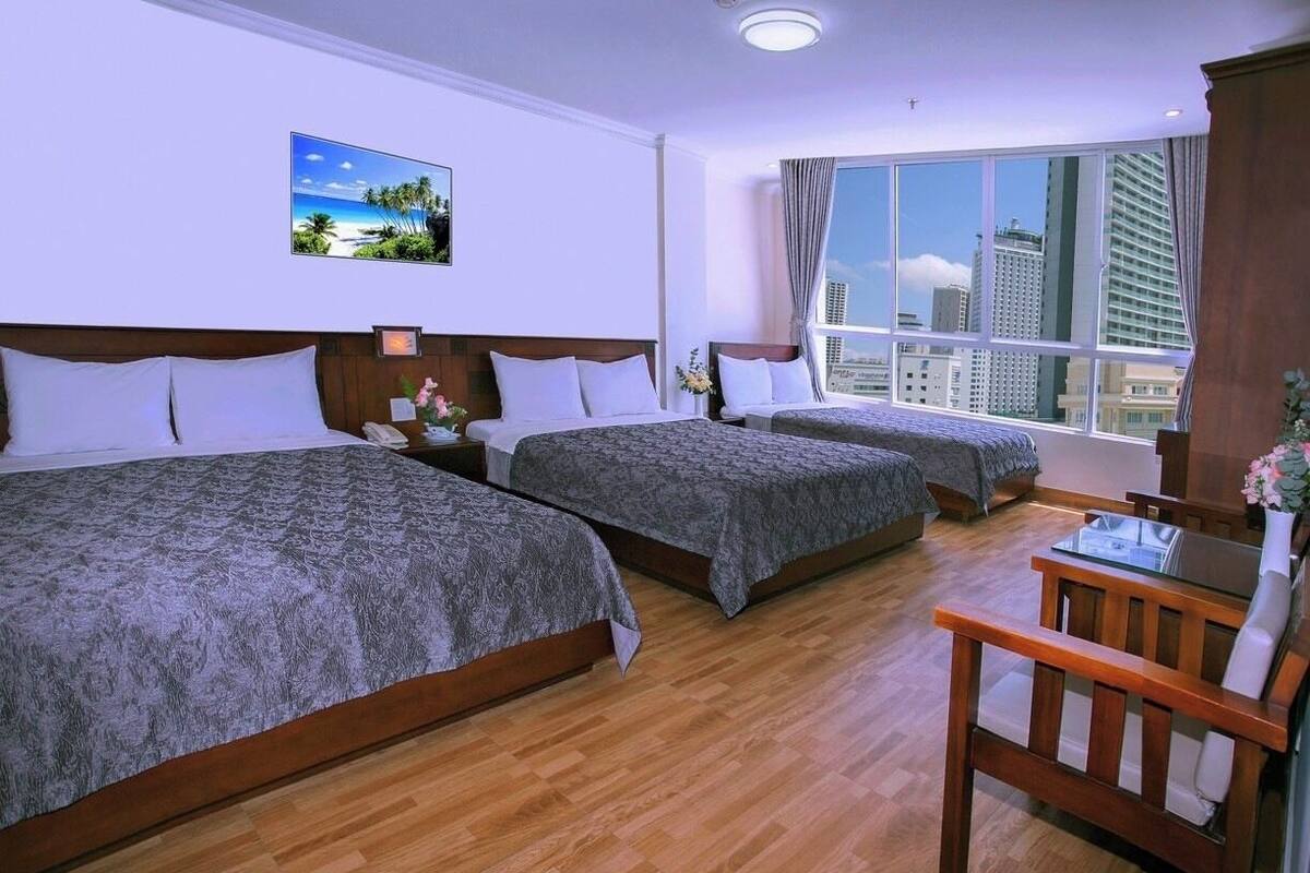 A hotel room with two beds, a wooden floor, a desk with a chair, and a large window with a city view.