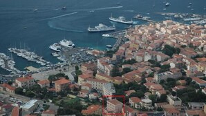 Vista aérea de un pueblo costero con un puerto deportivo, edificios con techos de tejas rojas y barcos.