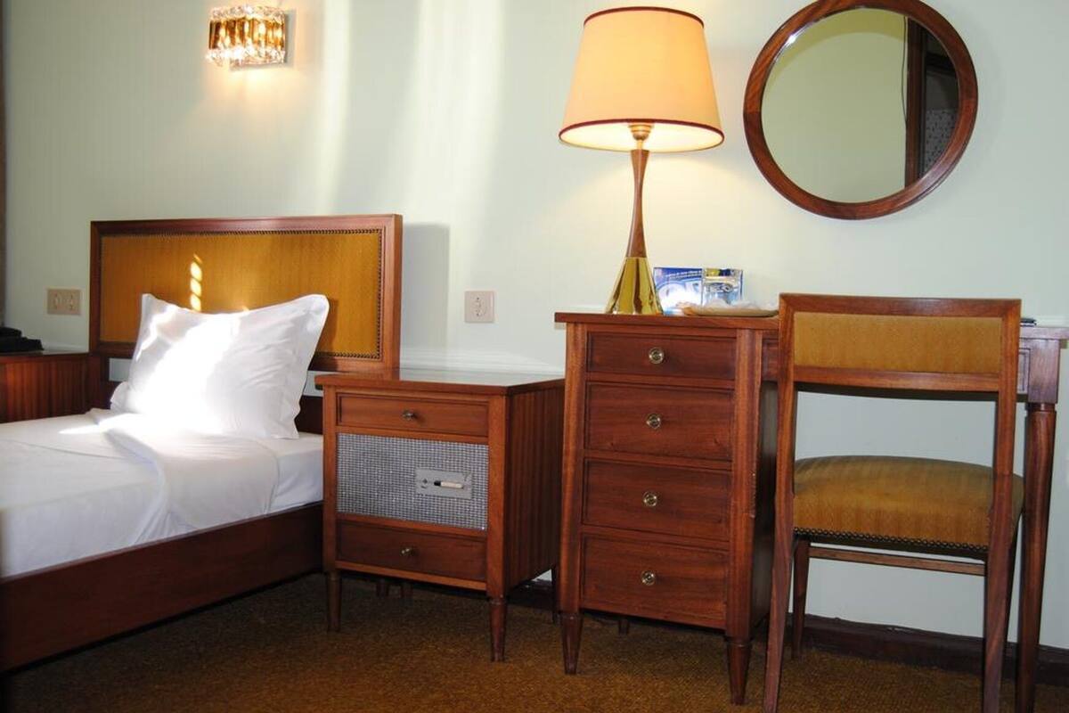 A hotel room with a bed, a wooden nightstand with drawers, a desk with a lamp, and a chair.