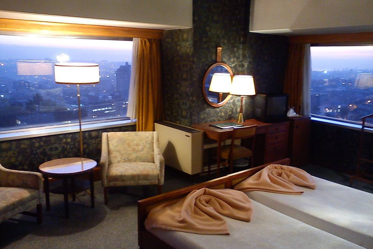 A hotel room with a bed, a desk, a chair, a small table, and a view of the city through large windows.