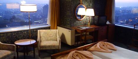 Habitación de hotel con cama, escritorio, silla, mesita y vistas a la ciudad a través de amplios ventanales.