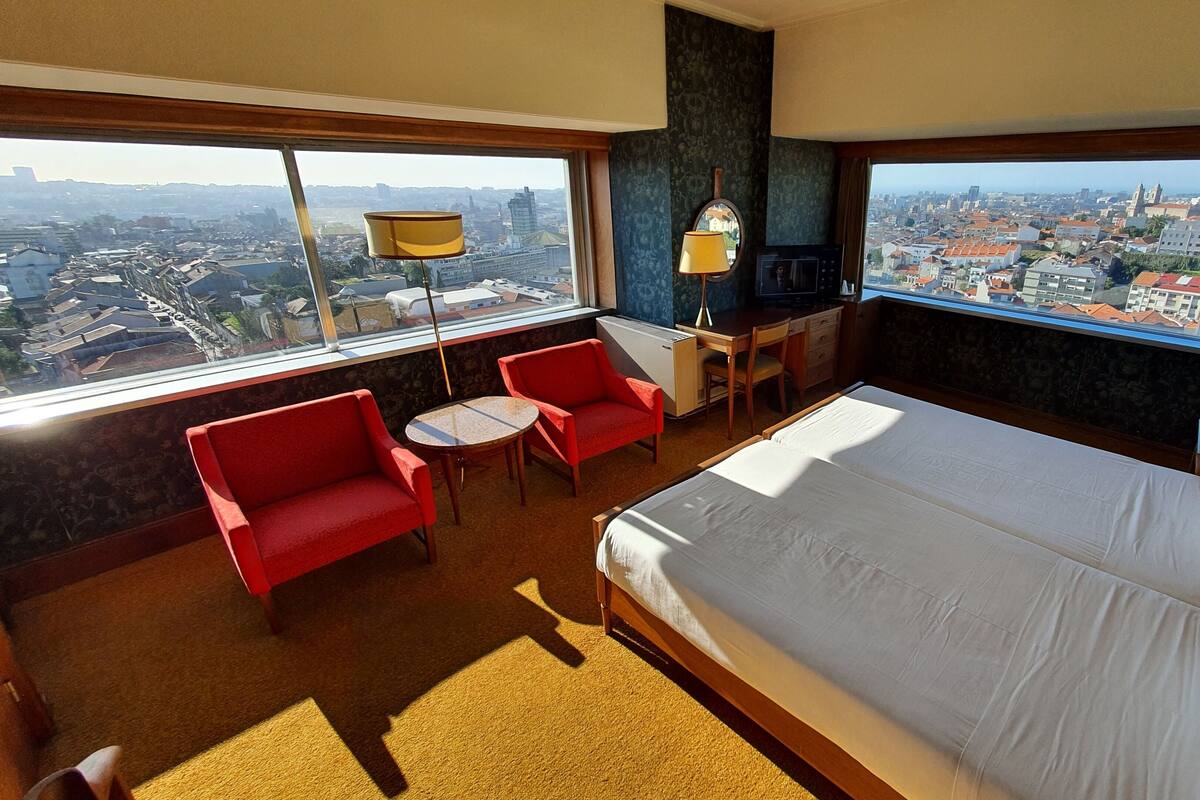 A hotel room with a large bed, two red chairs, a small table, a desk with a lamp, and a view of a cityscape through large windows.