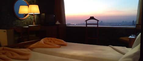 Une chambre d’hôtel avec deux lits, une télévision, une lampe et une vue sur les toits de la ville au coucher du soleil.