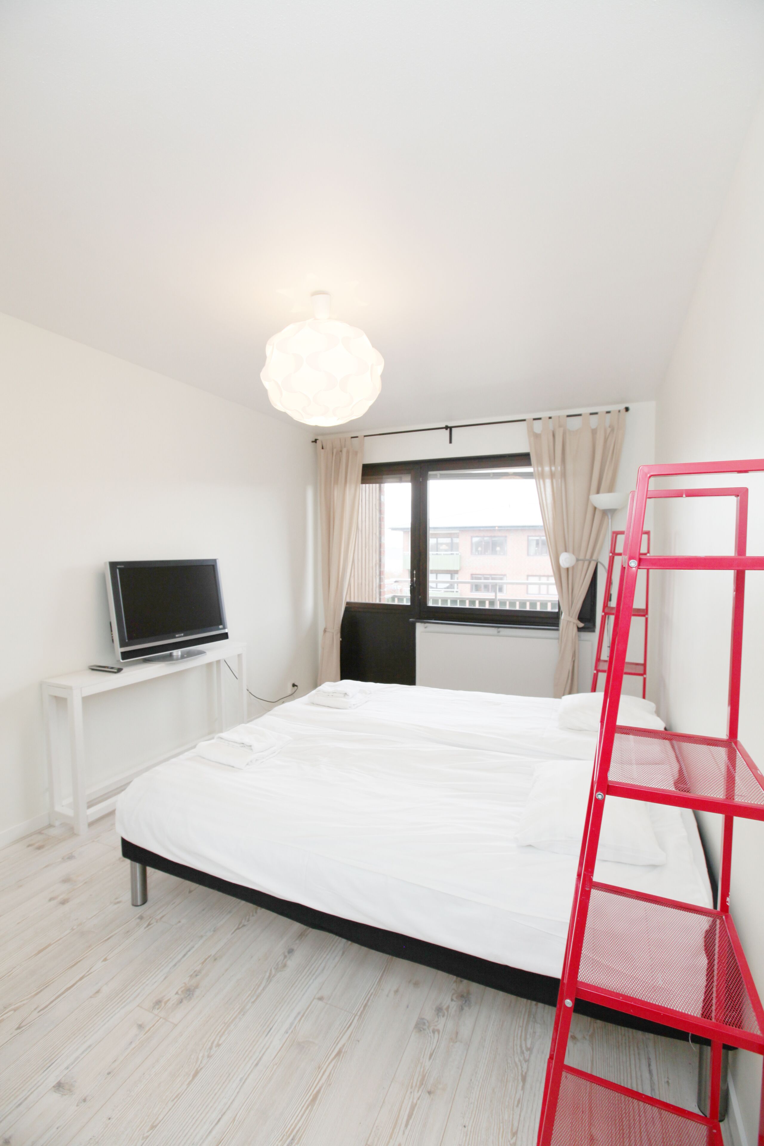 A room with a bed, a TV on a stand, a window with curtains, and a red ladder.