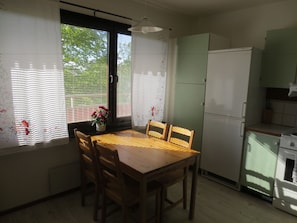 A kitchen with a wooden table and chairs, a refrigerator, and a window with blinds. - Fjordhotellet (Lysekil)