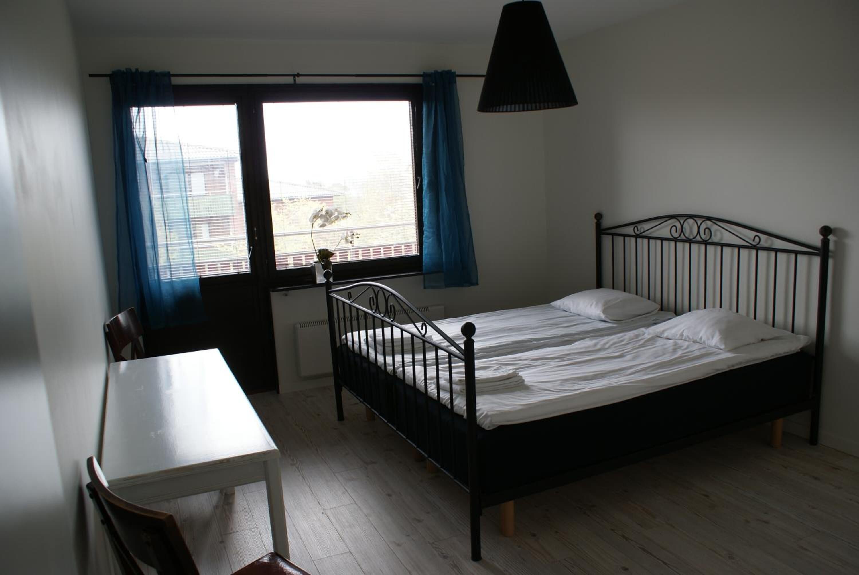 A bedroom with a bed, a desk, and a chair. There is a window with blue curtains and a view of buildings outside.