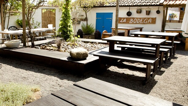 Un patio exterior con mesas y bancos de madera, una puerta azul con el rótulo "LA BODEGA" y un entorno de jardín rústico.