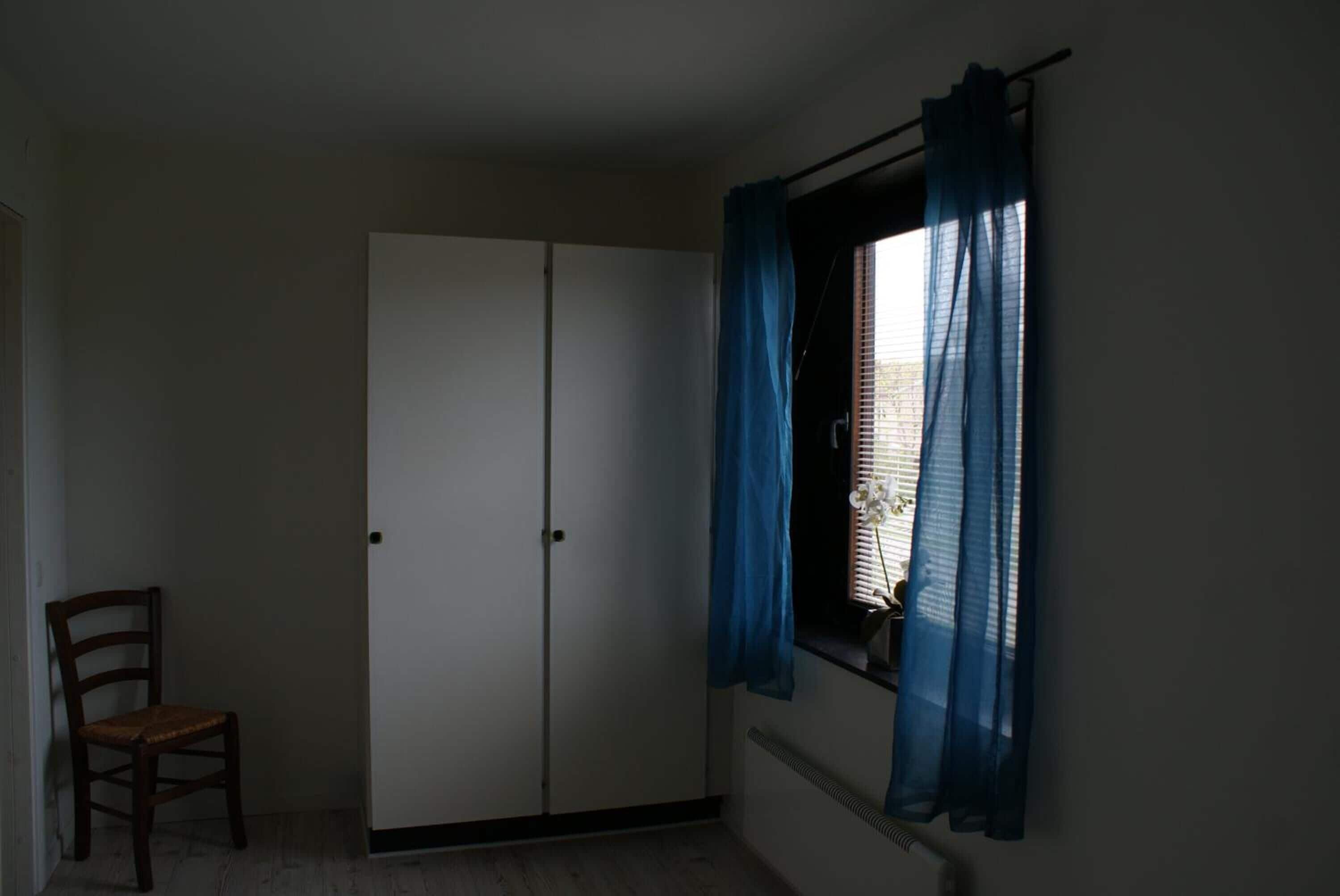 Una habitación con armario, una silla y ventana con cortinas azules.