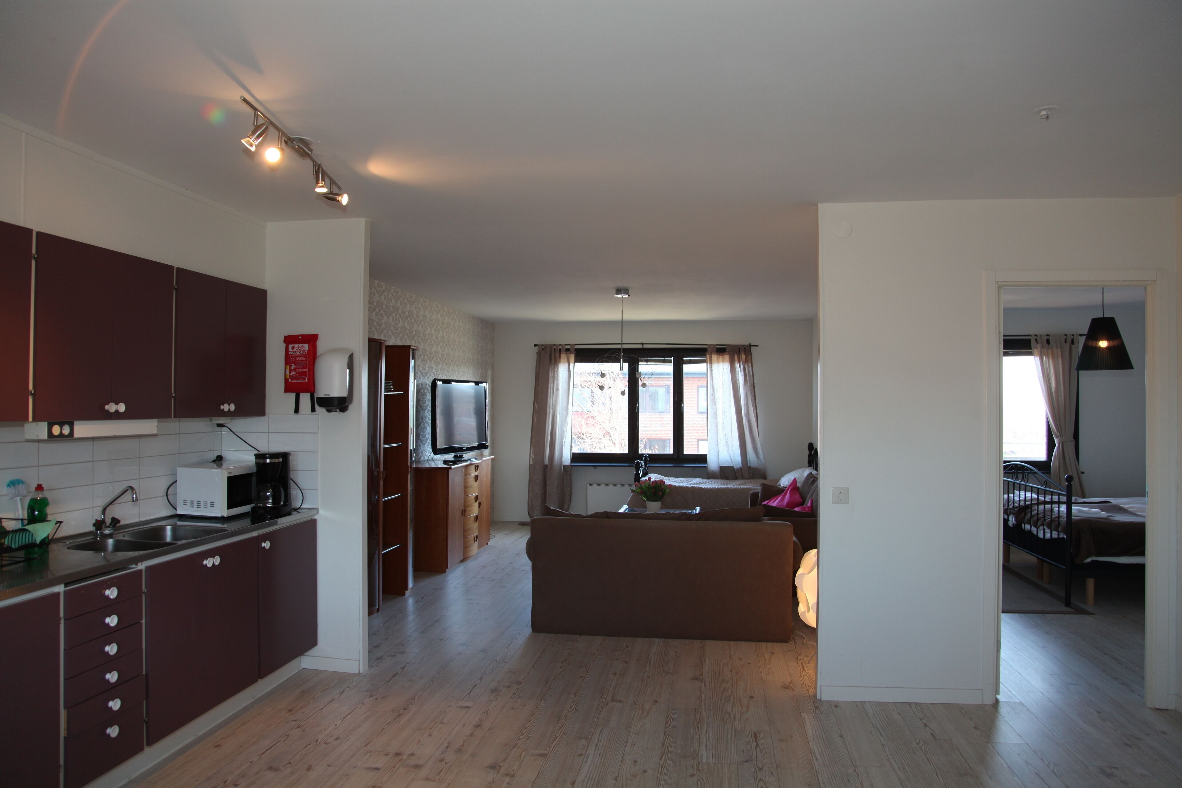 A modern kitchen with dark cabinets, a built-in microwave, and a coffee maker. There is a dining area with a large window and curtains, and a separate bedroom with a bed and a nightstand.