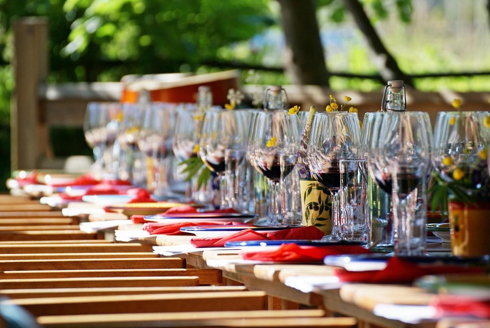 A row of tables with clear glassware and red napkins, set outdoors with a wooden fence in the background.
