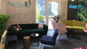 A hotel lobby with a green sofa, a blue chair, and a yellow sofa. There are plants, a neon sign, and a door with a window. - Ekho Hotel Grenoble Nord Saint Egrève (Saint-Egreve)