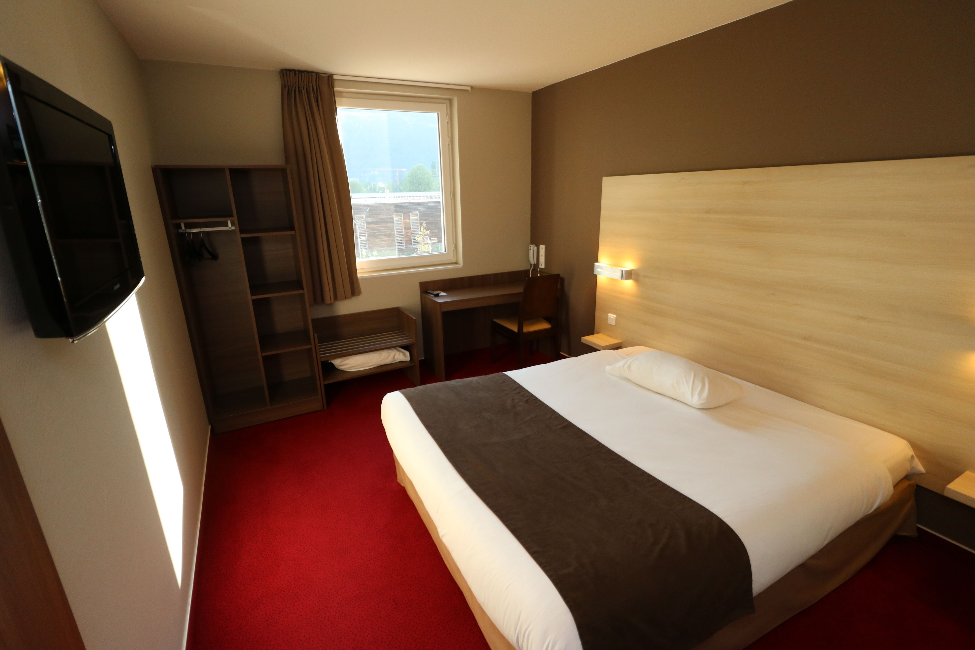 A hotel room with a large bed, a desk, a chair, a TV, and a window with curtains.
