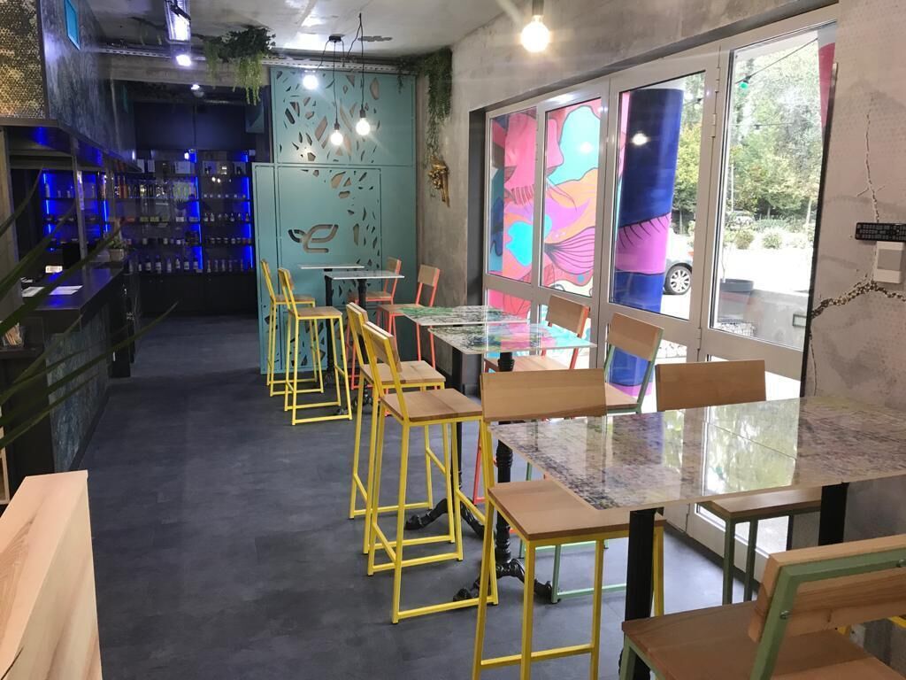 A restaurant with a bar area, tables with yellow chairs, and large windows with colorful artwork.