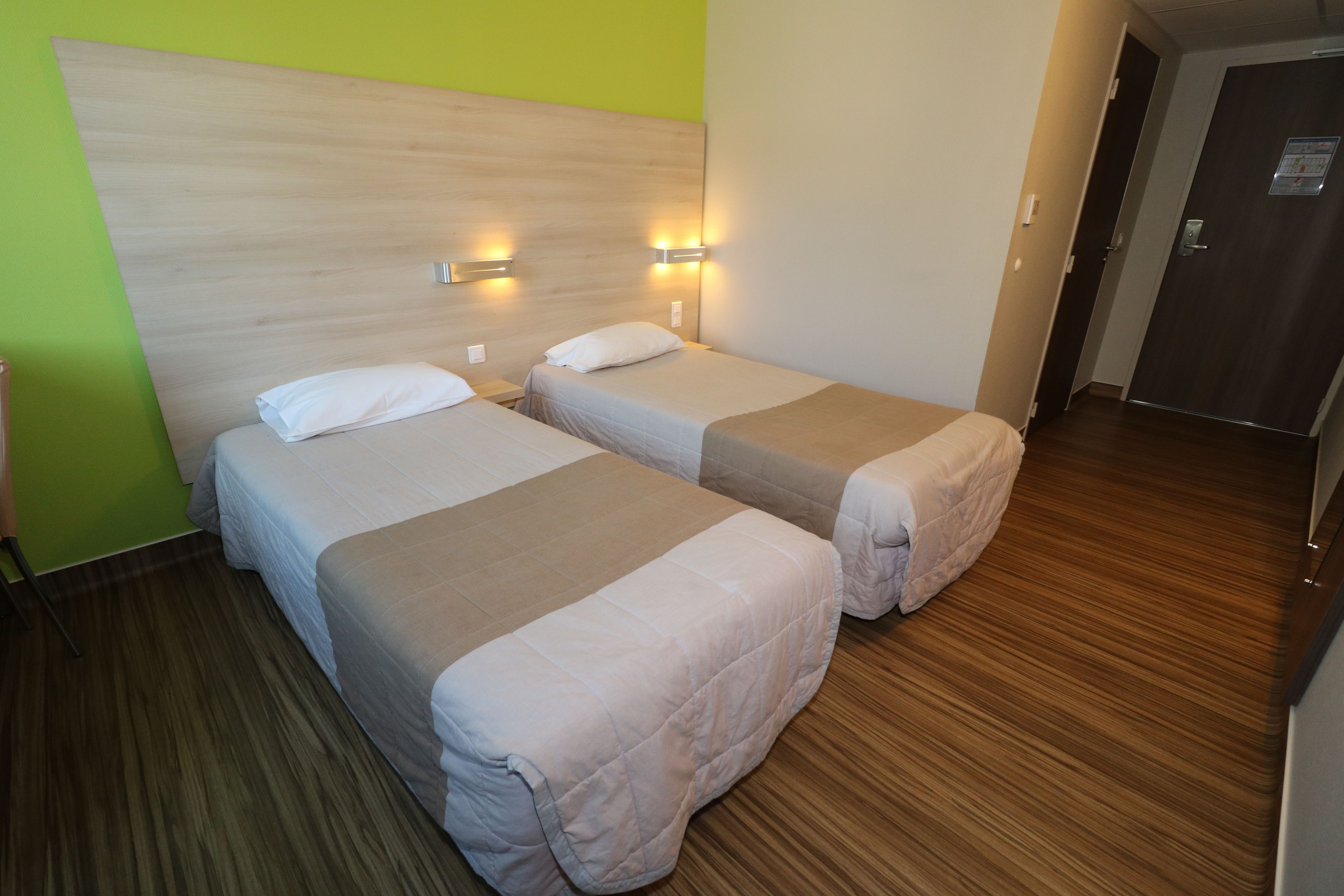 A hotel room with two beds, wooden headboard, and nightstands with wall-mounted lamps.