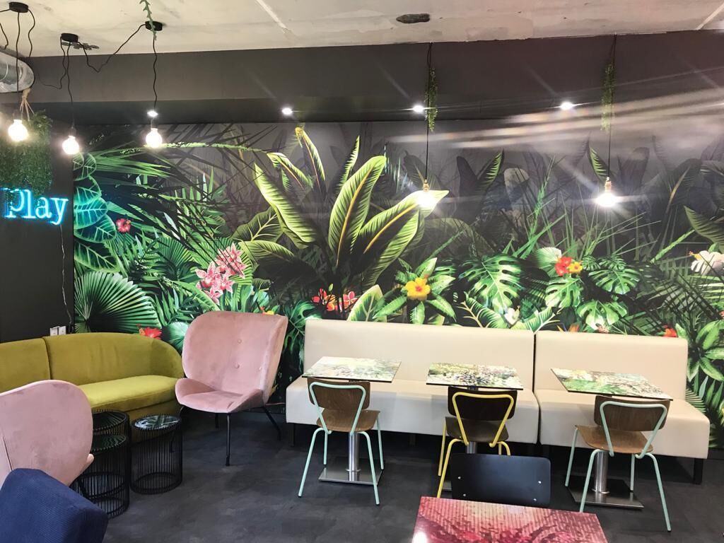 A room with a mural of tropical plants, a neon 'Play' sign, a yellow sofa, pink chairs, and small tables with chairs.