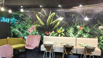 A room with a mural of tropical plants, a neon 'Play' sign, a yellow sofa, pink chairs, and small tables with chairs.