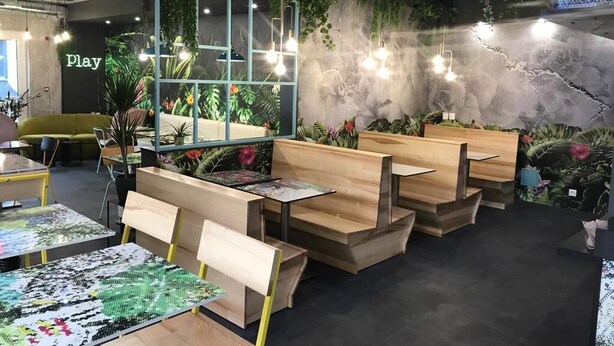 A modern cafe with wooden benches and tables, green plants, and a 'play' sign on the wall. - Ekho Hotel Grenoble Nord Saint Egrève (Saint-Egreve)