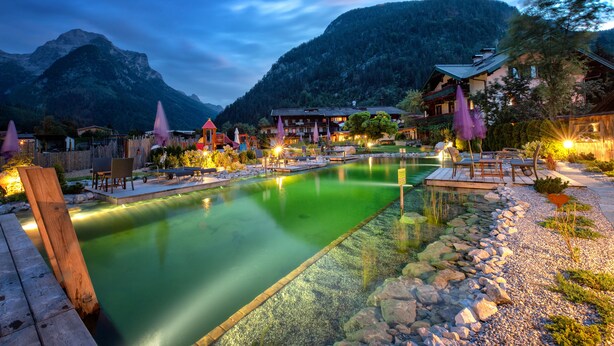 Un área de piscina tranquila al aire libre con cubierta de madera, rodeada de rocas y vegetación, con montañas y edificios tradicionales al fondo.