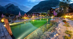 A serene outdoor pool area with a wooden deck, surrounded by rocks and greenery, set against a backdrop of mountains and traditional buildings. - Mühlpointhof (Lofer)