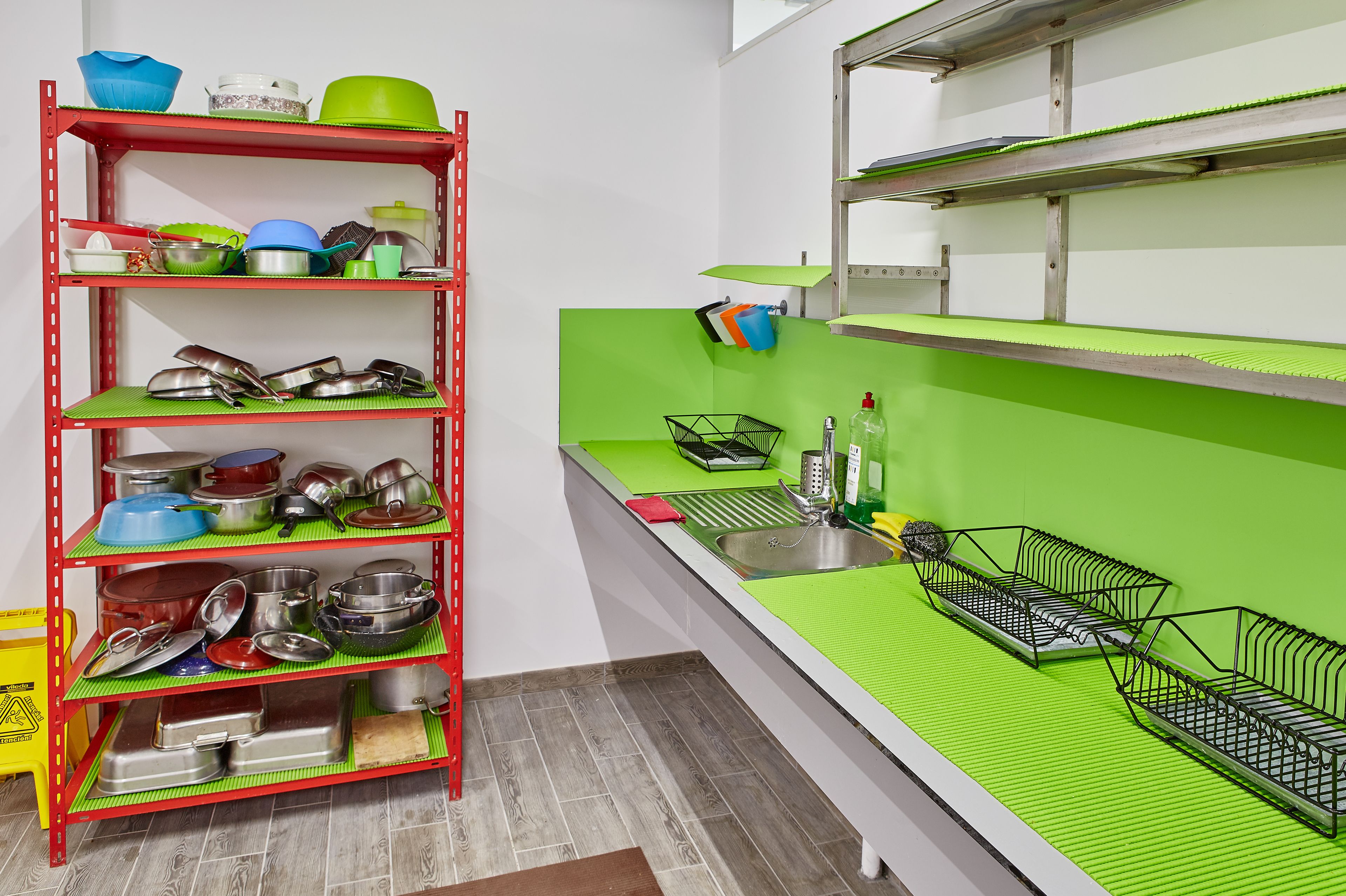 A kitchen with a red metal shelving unit filled with pots and pans, a green countertop, and a sink with a green mat.