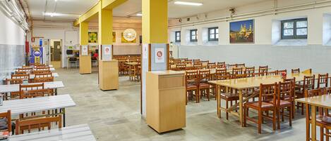 Cafetería con varias mesas y sillas, un pilar central y una pared con ventanas y un cartel.