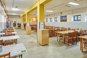 A cafeteria with multiple tables and chairs, a central pillar, and a wall with windows and a poster. - Albergue Seminario Menor - Hostel (Santiago de Compostela)
