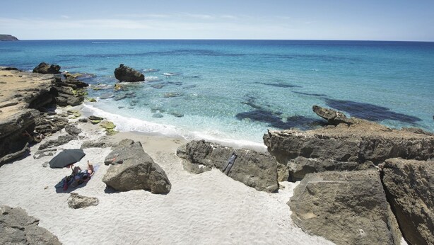 Una playa rocosa con agua turquesa cristalina, una zona arenosa con una persona bajo una sombrilla y grandes rocas.