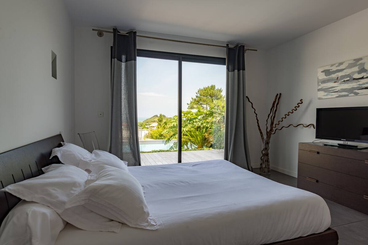 A bedroom with a large bed, a TV, and a view of a swimming pool and greenery.
