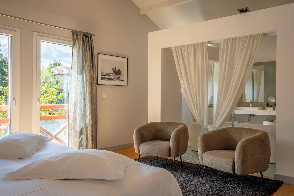 A bedroom with a bed, two armchairs, a black rug, a large window with curtains, and a bathroom visible through a glass door.