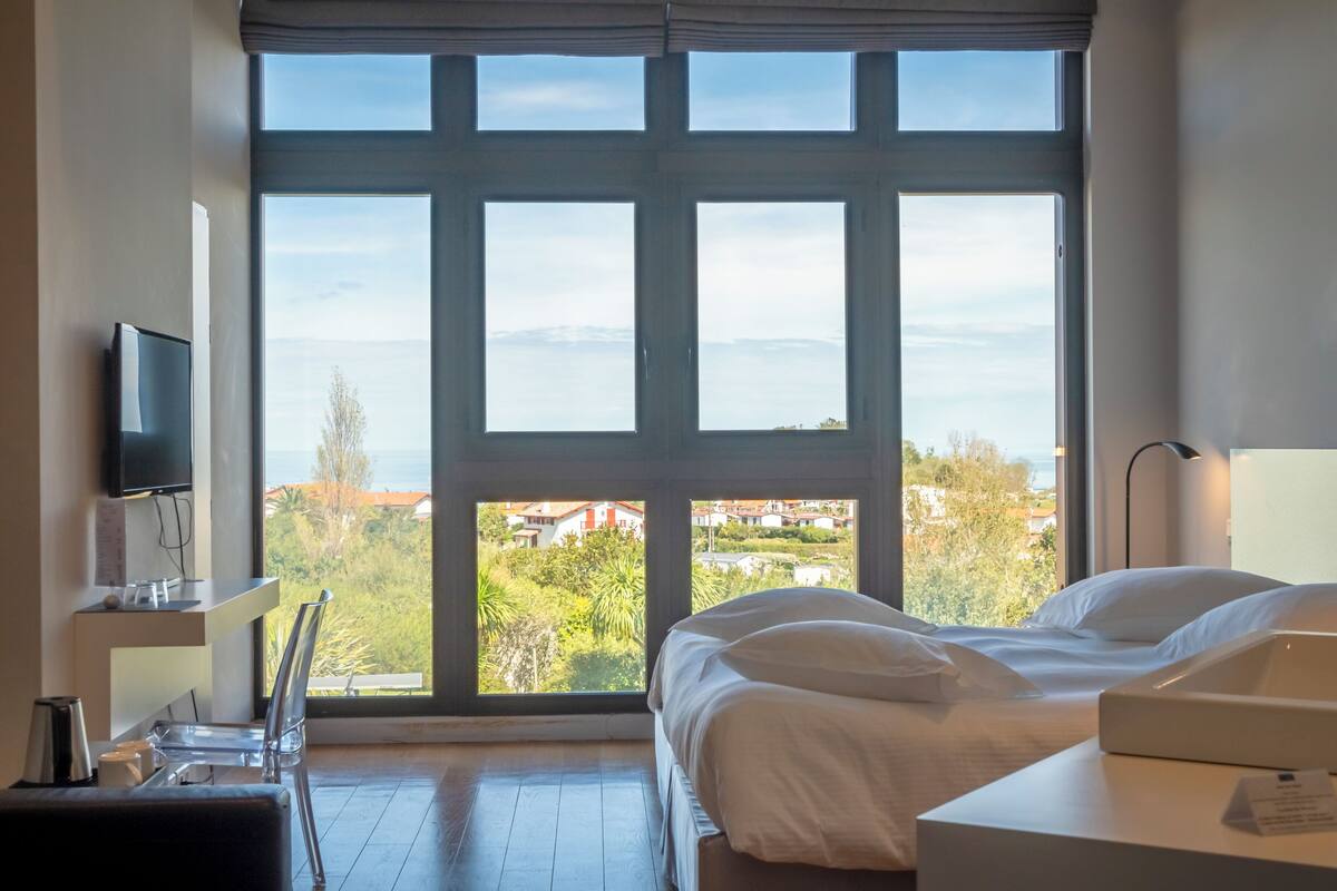 A hotel room with a large window, a bed, a desk with a chair, and a TV.