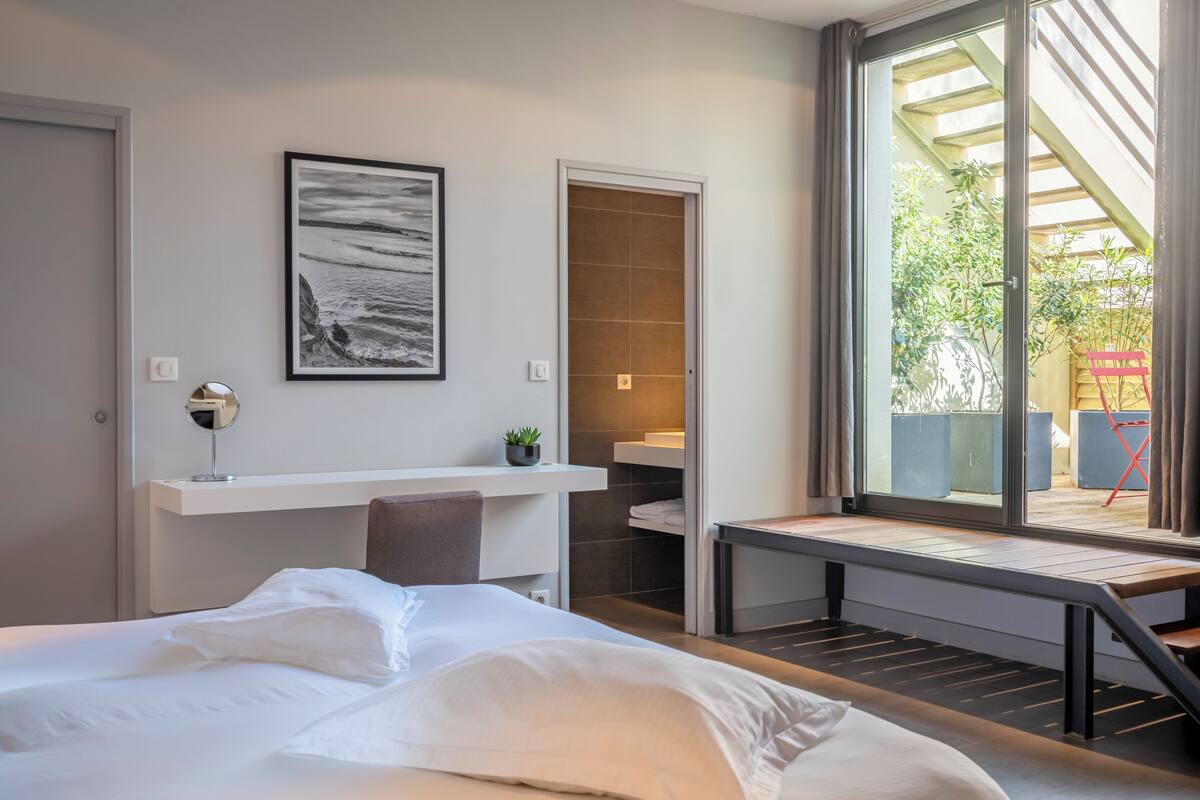A modern hotel room with a bed, a desk, a small plant, a bathroom, and a view of the outdoors.