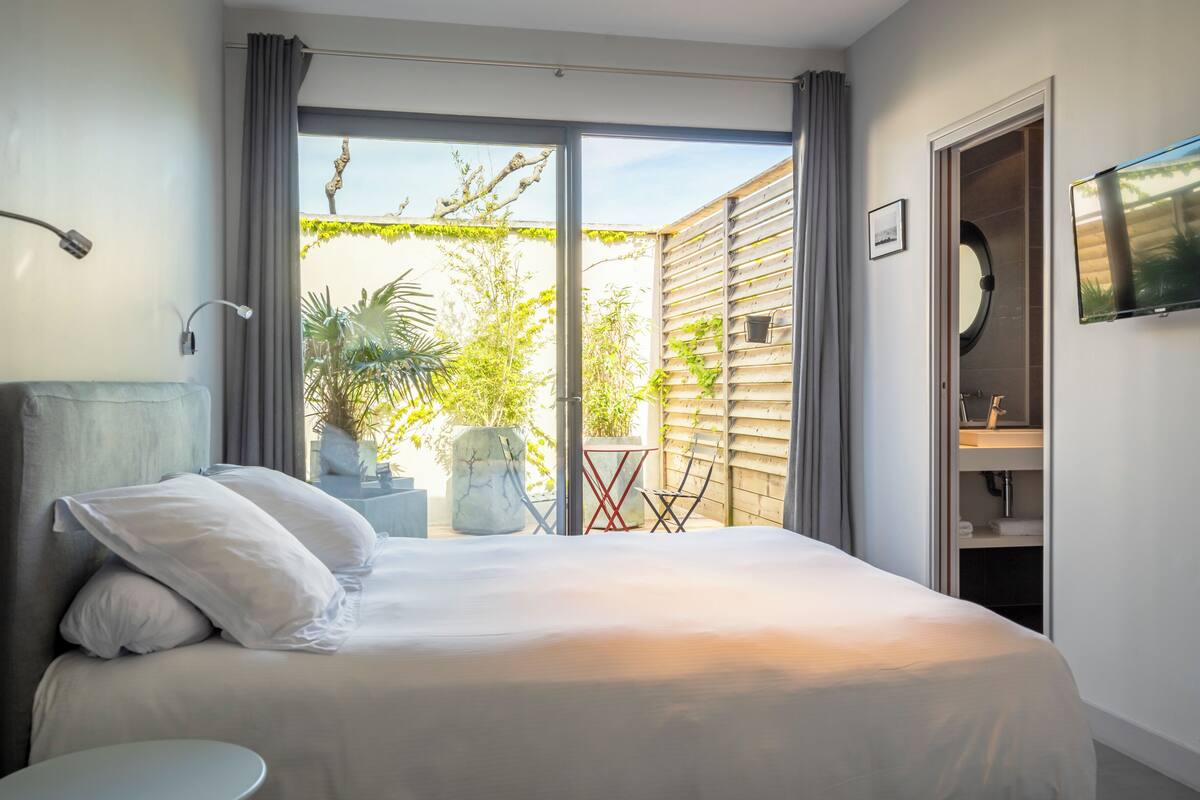 A bedroom with a bed, a TV, a window with curtains, a plant, and a small table.