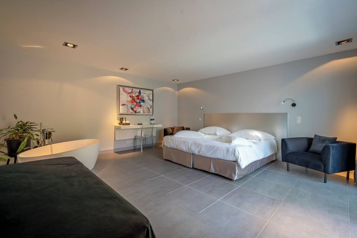 A modern bedroom with a large bed, a desk, a chair, and a bathtub.