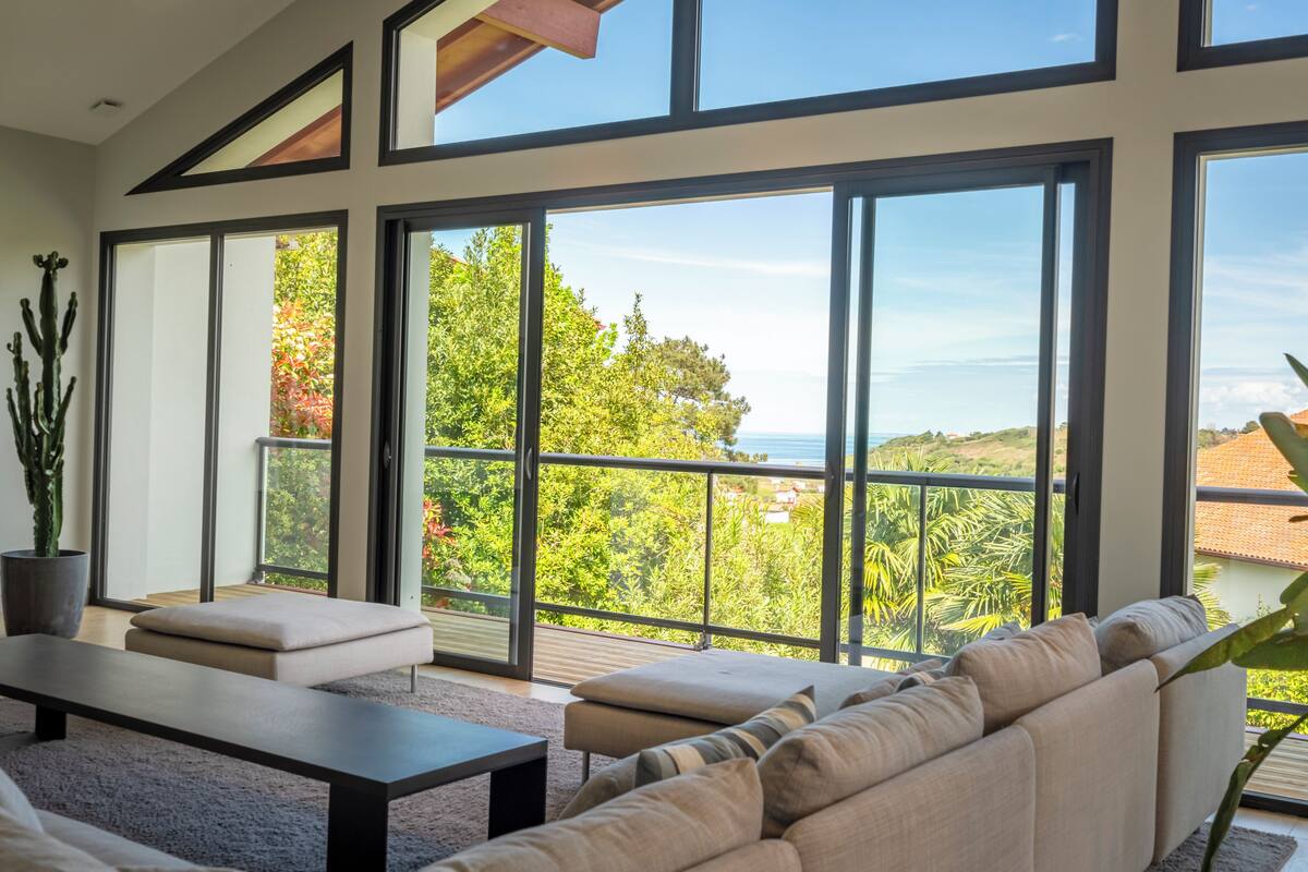 A modern living room with a large sliding glass door leading to a balcony with a view of greenery and the sea.