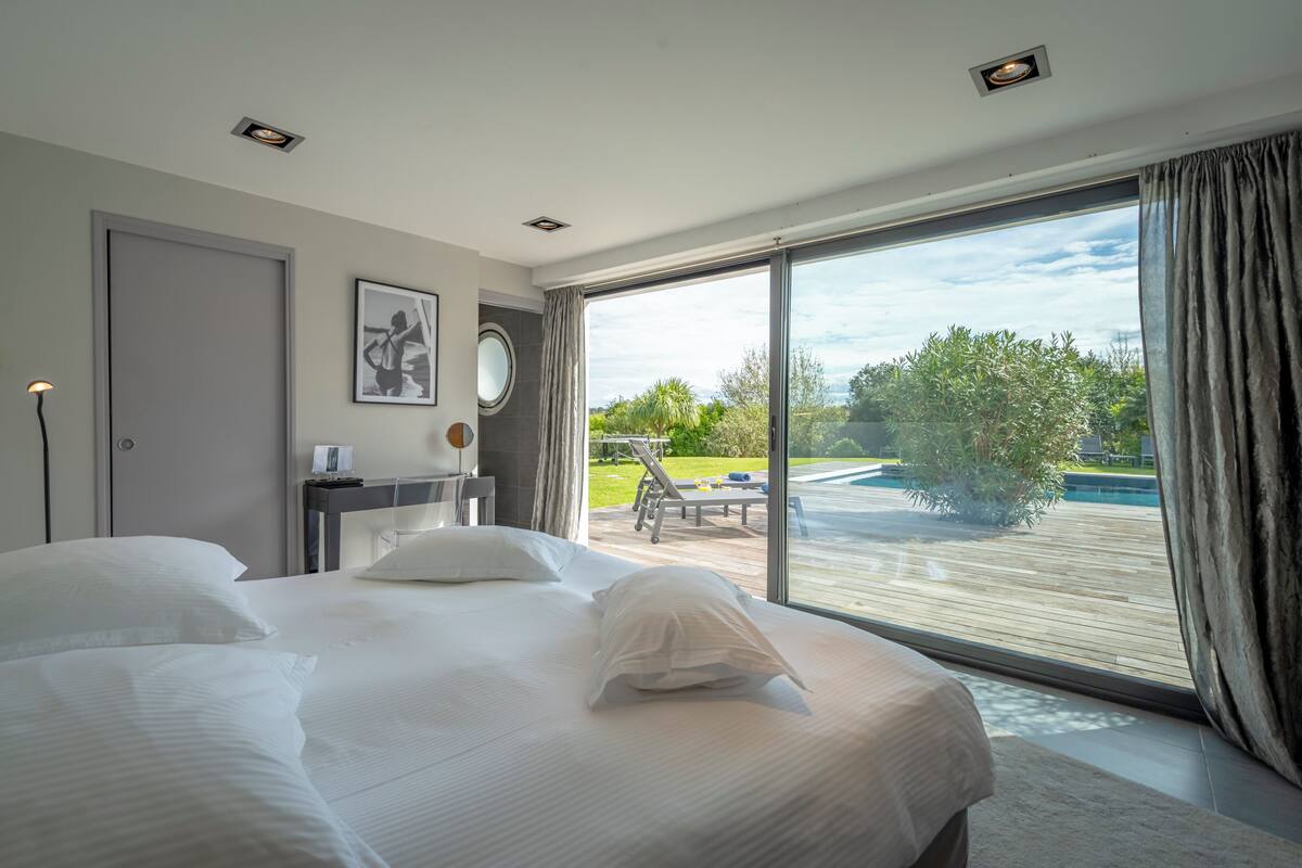A hotel room with a large bed, a desk, a chair, a round mirror, and a view of a swimming pool and greenery.