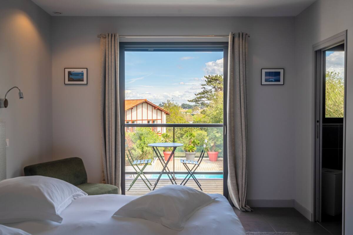 A hotel room with a bed, a chair, a small table, and a balcony with potted plants and a view of the outside.