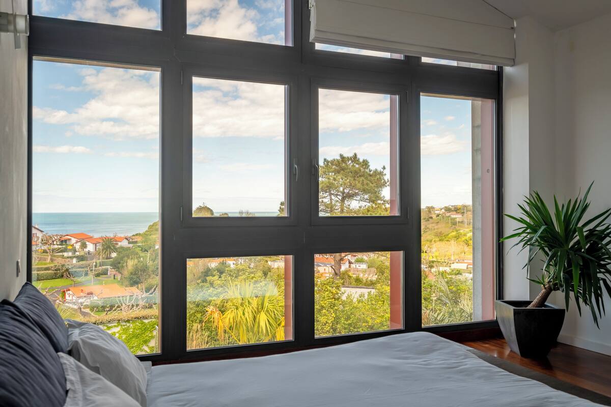 A bedroom with a large window offering a view of a coastal town and the sea.