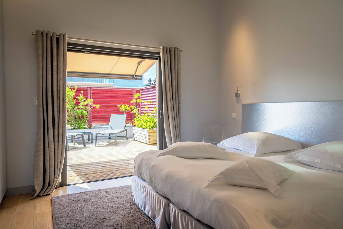 A bedroom with a bed, a nightstand, and a view of an outdoor patio with a red fence and greenery.