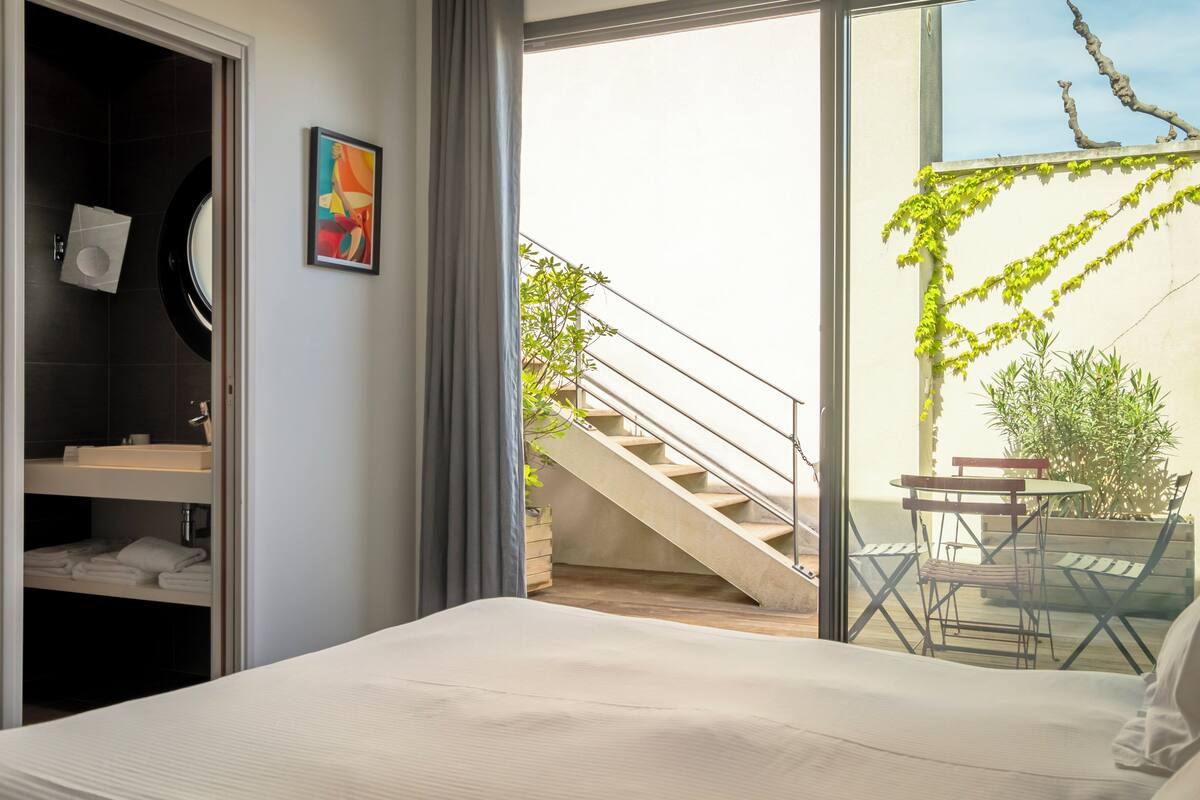 A hotel room with a bed, a bathroom with a towel rack, a staircase leading to an outdoor area with a table and chairs, and a view of greenery.