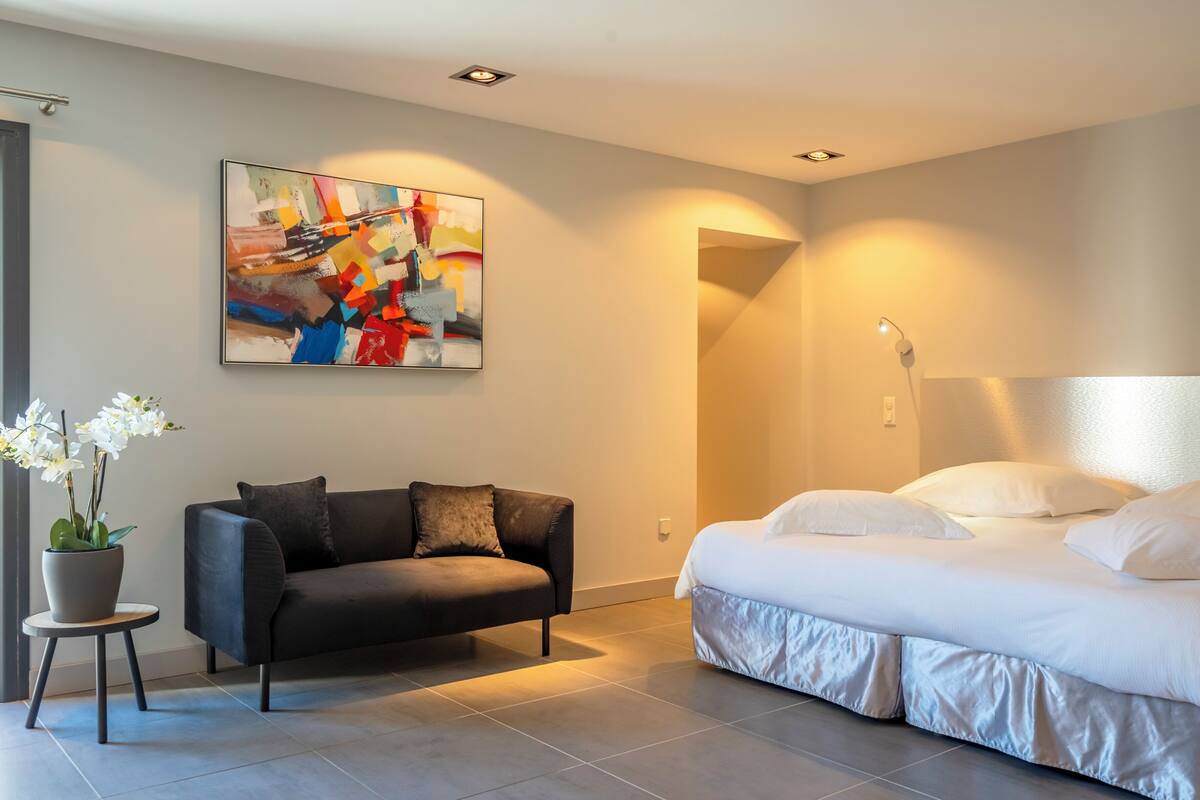 A modern bedroom with a large bed, a dark sofa, a small table with a plant, and a colorful abstract painting on the wall.