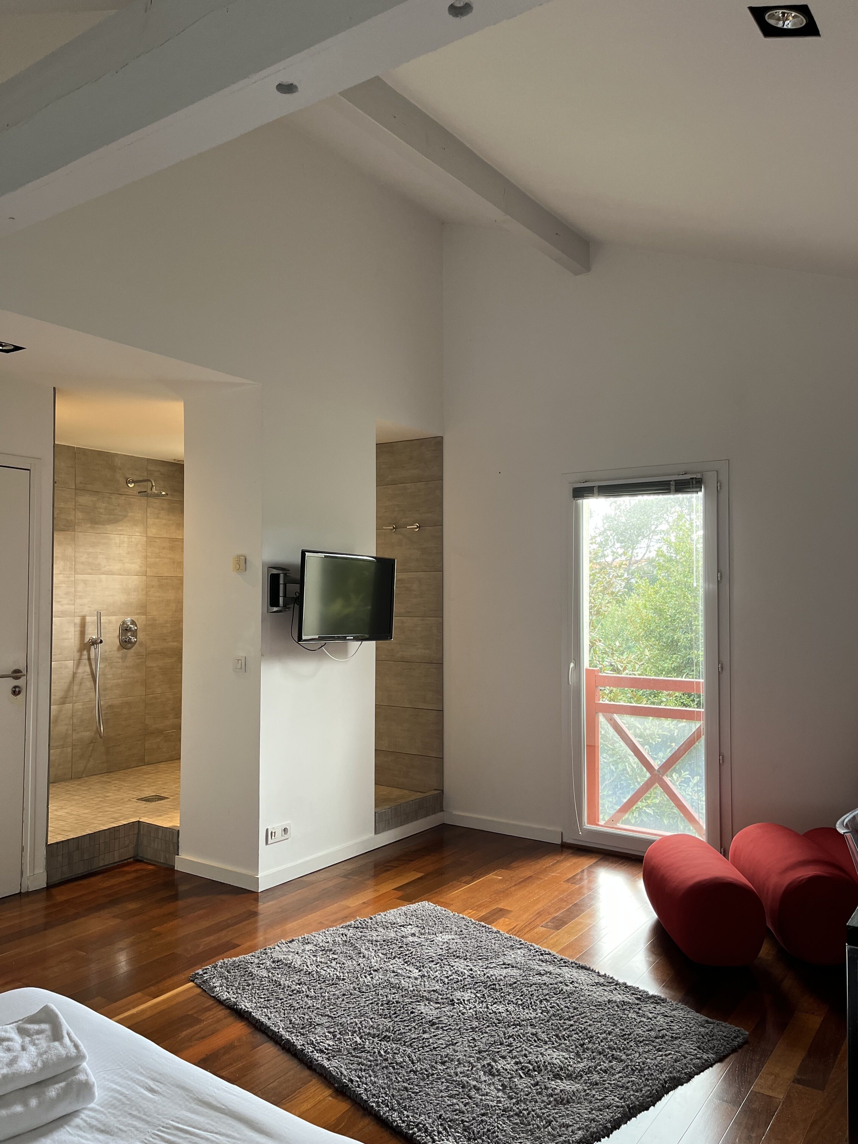 A modern bathroom with a shower, a toilet, and a sink. There is a television mounted on the wall and a window with a view of greenery outside.