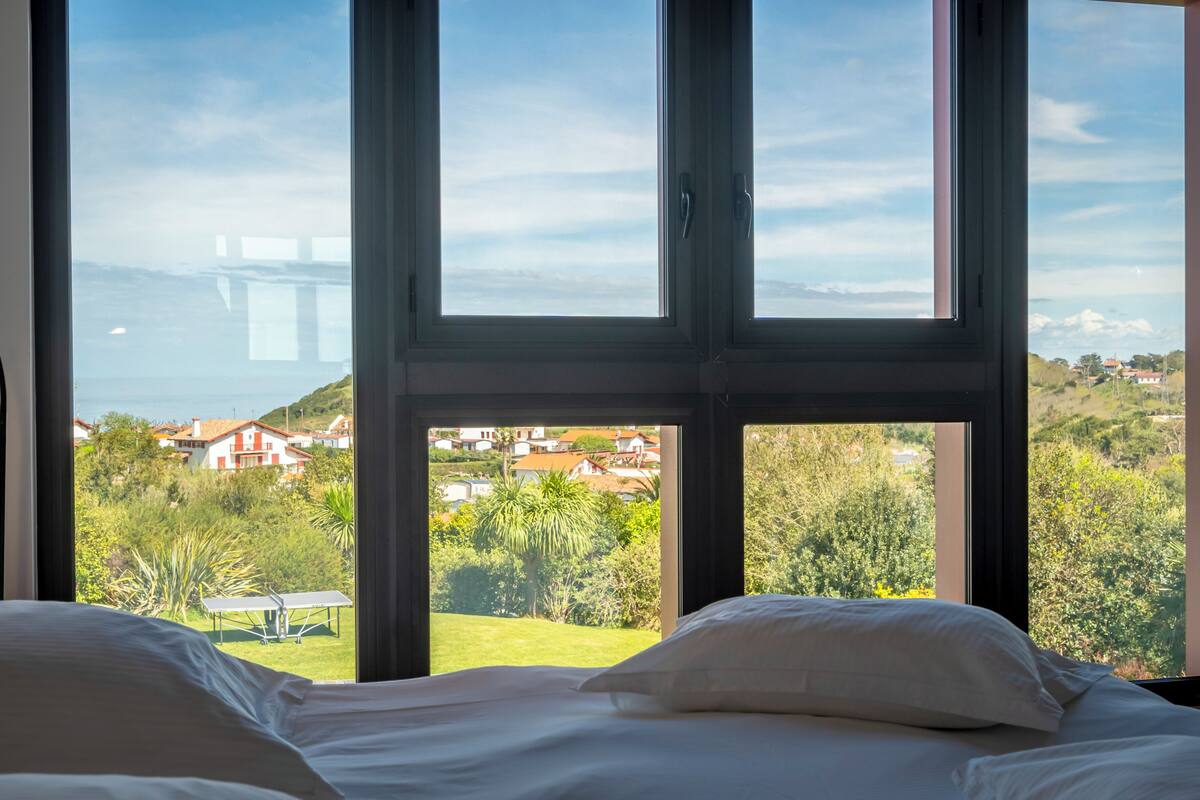 A bedroom with a large window offering a view of a coastal town and greenery.