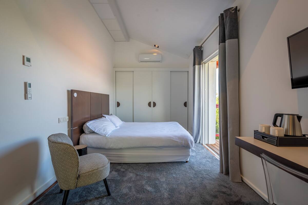 A hotel room with a bed, a chair, a desk with a kettle, and a TV.