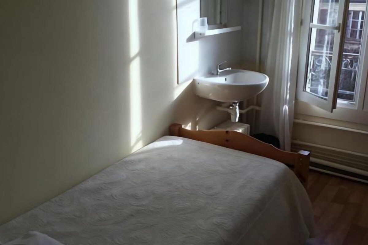 Un dormitorio pequeño y ordenado con una cama individual, una mesita de noche blanca, un lavamanos blanco empotrado y una ventana con cortinas.