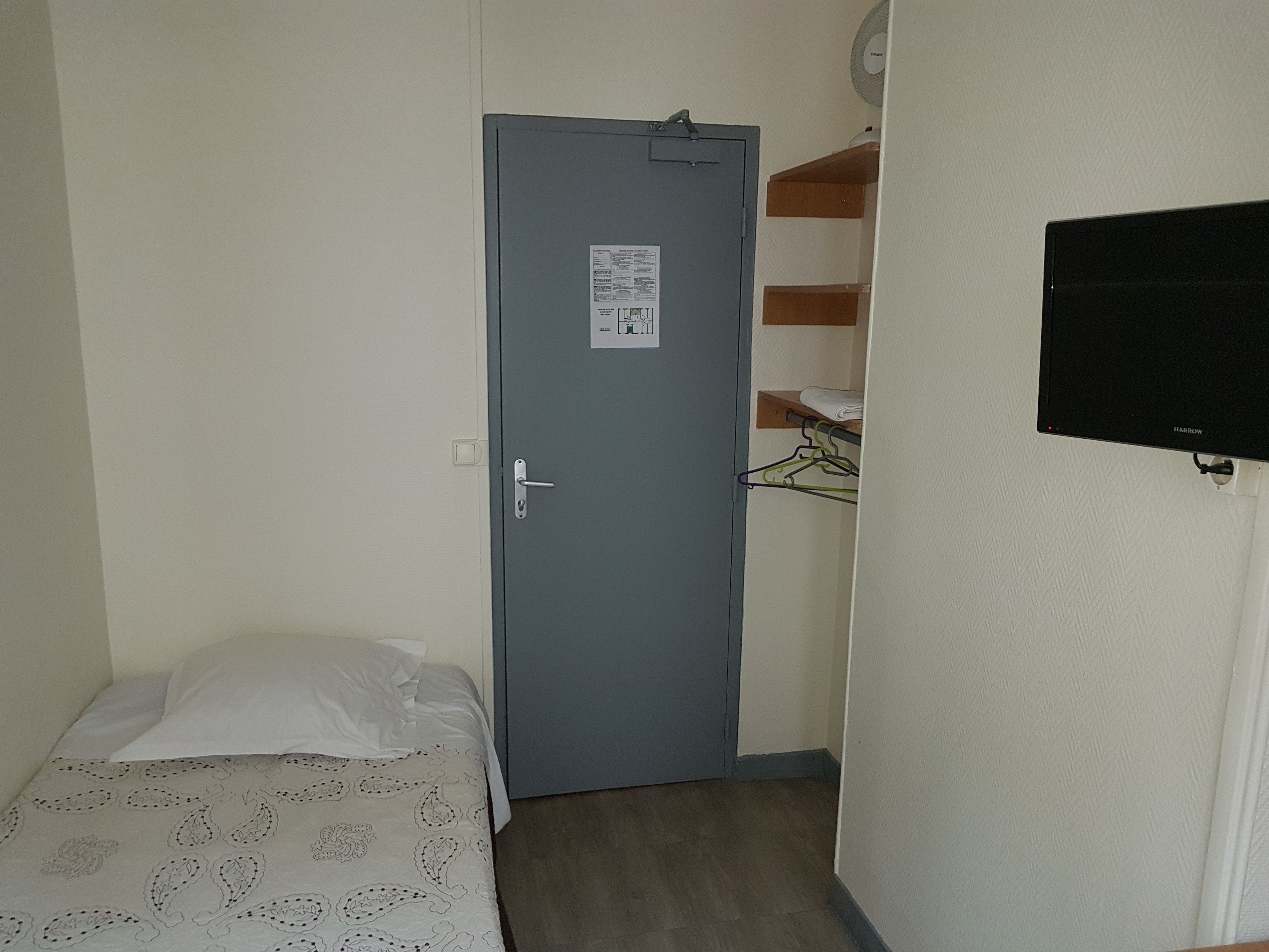 Ein kleines Zimmer mit einem Bett, einer grauen Tür mit einem Schild, einem hölzernen Regal und einem an der Wand befestigten Fernseher.