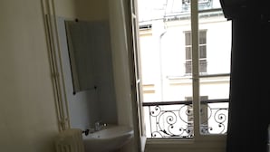 A room with a bed, a window with a view of buildings, a radiator, and a small sink. - Au Royal Mad Hotel (Paris)