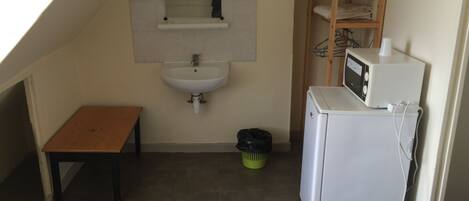 Una habitación pequeña y estrecha con un banco de madera, un lavabo, un espejo, un microondas y una cama con colcha estampada.