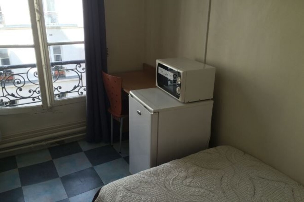 A small room with a bed, a desk with a microwave, a chair, and a window with a view of buildings.