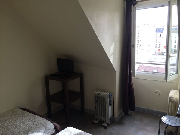 Una habitación con una cama, una mesita con una computadora, un radiador y una ventana que da a vistas de edificios.